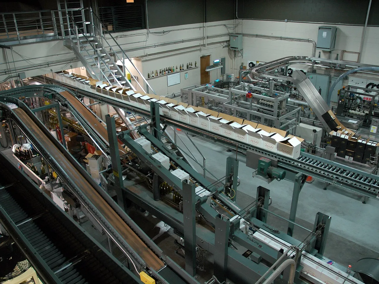 Elevated view of an industrial bottling line with conveyor belts, packaging cartons, and pump-driven fluid transfer equipment.